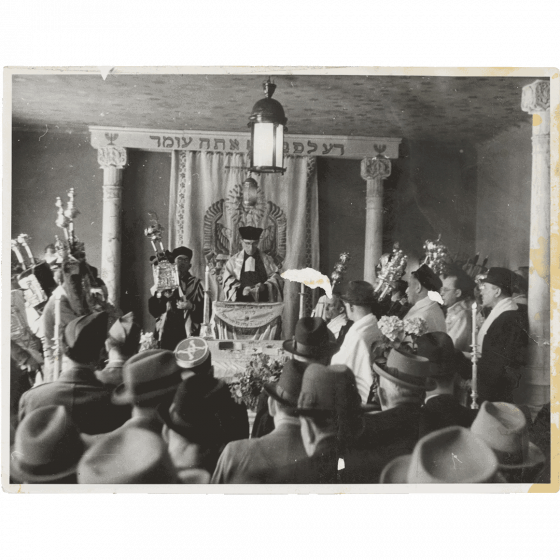 Black and white photograph of a synagogue, in the center a rabbi, around him men with Judaica 