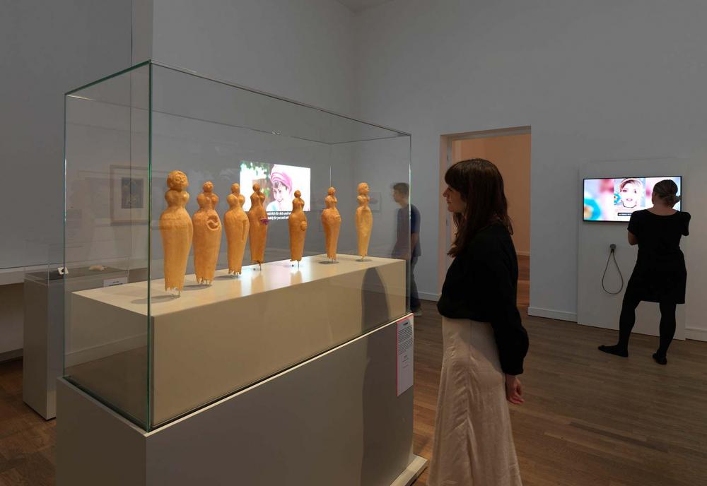 Ausstellungsansicht, eine Frau steht vor einer Vitrine, in der sich orange Figuren befinden.