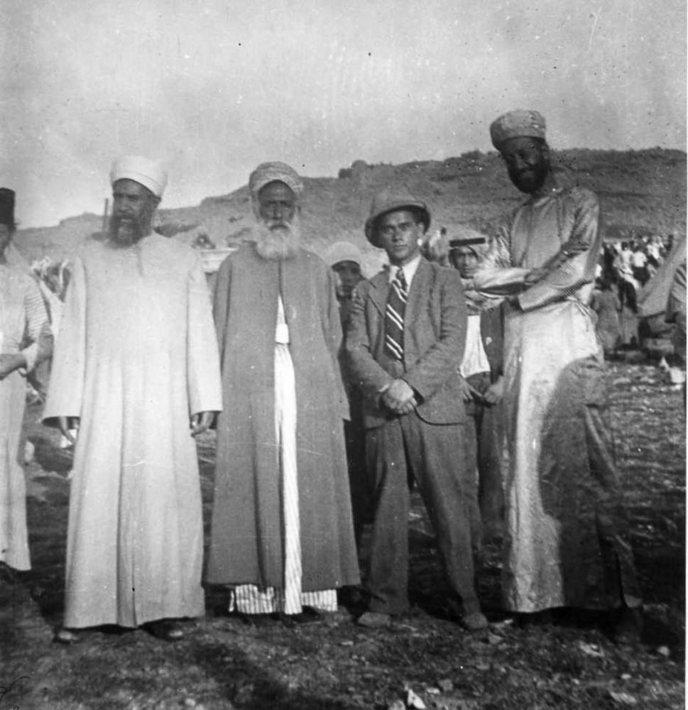 Black-and-white photo of four men in a desert landscape: one in a suit, tie, and hat; the other three with long beards and robes