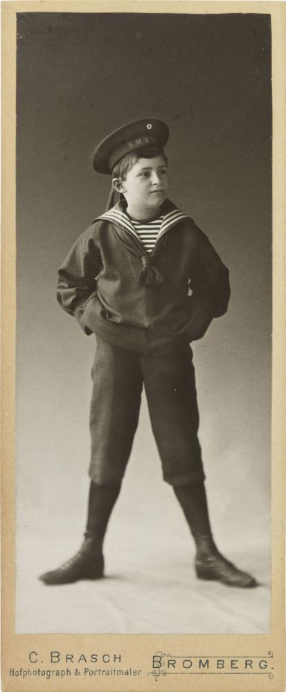Black-and-white photograph: boy in a sailor costume standing with legs apart and arms akimbo; studio portrait