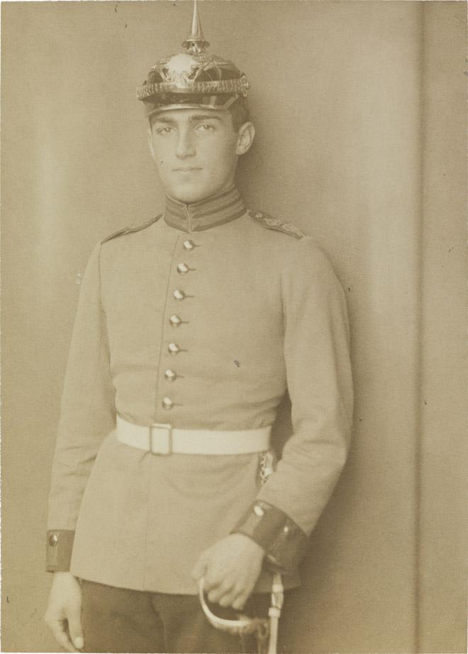 Black-and-white photograph: Young man in soldier’s uniform with white belt, spiked helmet, and sword, studio portrait