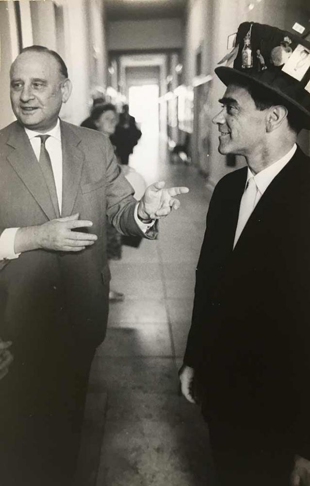 Black-and-white photo of a younger man with a decorated top hat and an older man pointing at him