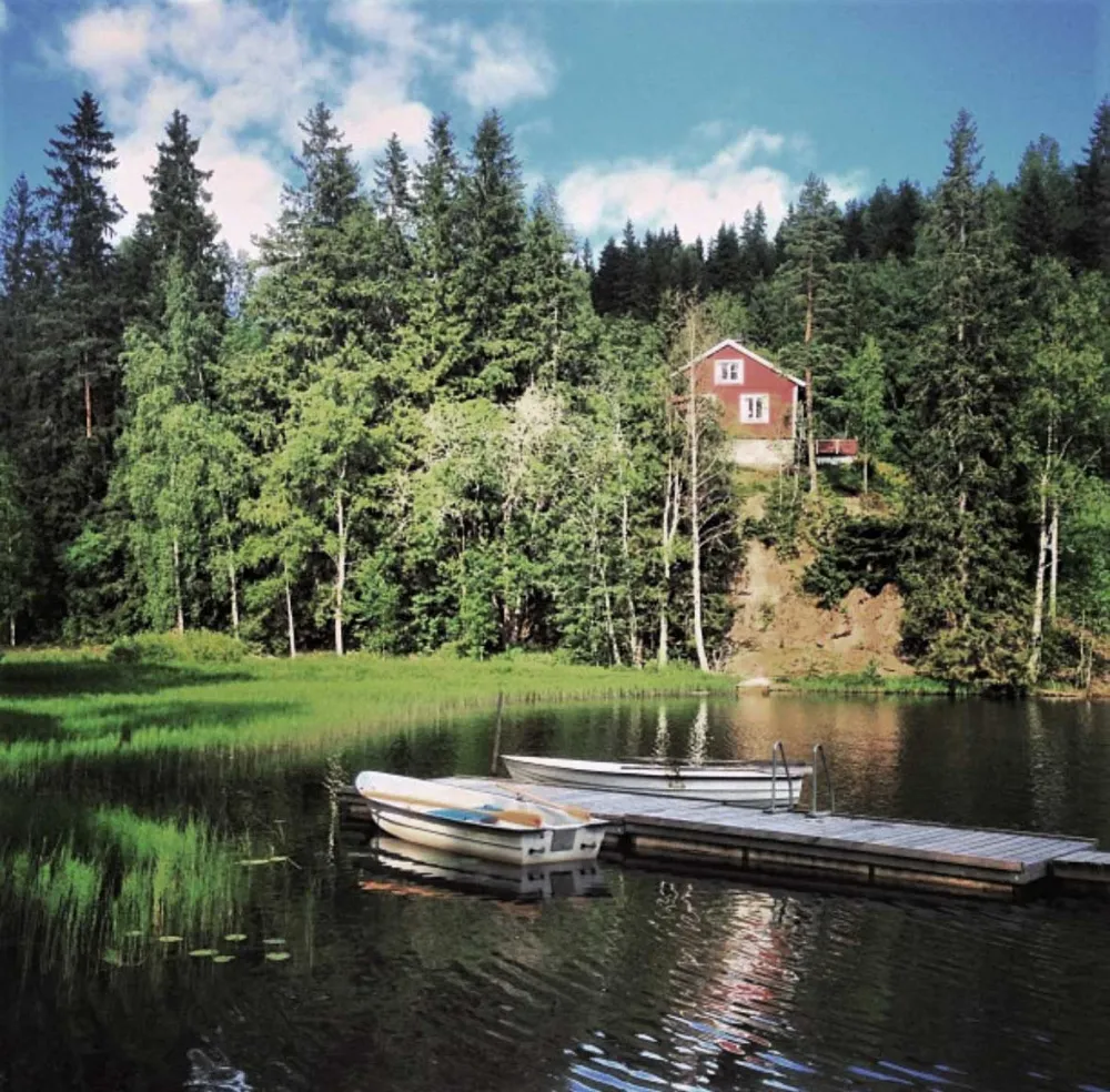 Im Vordergrund ein See mit Steg und zwei Booten. Mittel- und Hintergrund ein Wald, in dem idyllisch ein rotes Häuschen mit weißen versteckt liegt.