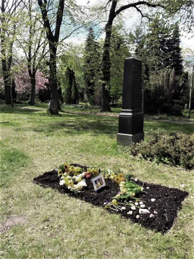 Ein frisches Grab, auf dem Blumen, kleine Steine liegen und ein gerahmtes Foto steht.
