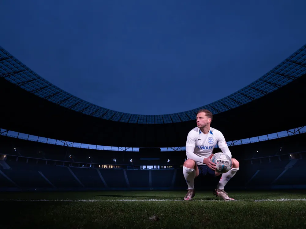 Ein Fußballspieler hockt im nächtlichen Olympiastadion mit einem Ball in der Hand und hat Kopf und Blick nach rechts gewendet.