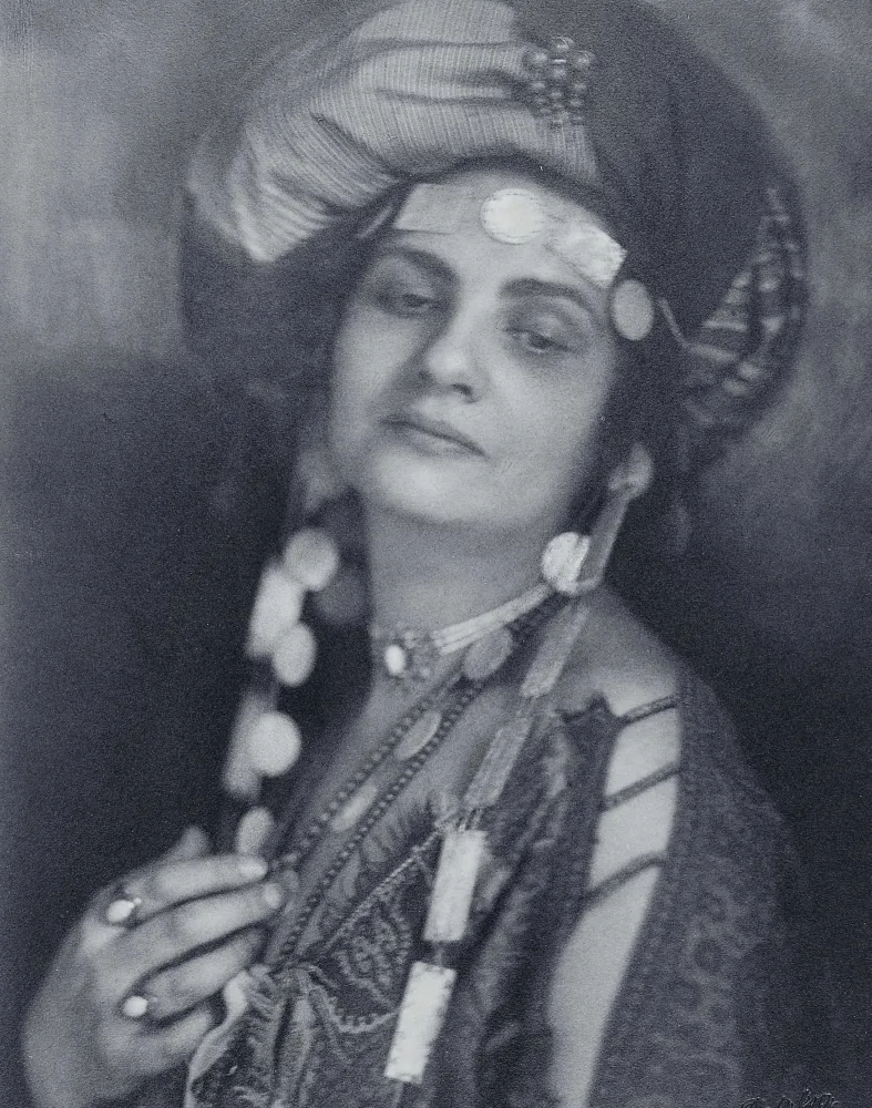 Die sepaifarbene Fotografie zeigt eine Frau mit Turban und einer Tunika und auffälligem Schmuck. Ihr Oberkörper und Gesicht sind zur Kamera gewendet aber ihr Blick geht Richtung Boden.
