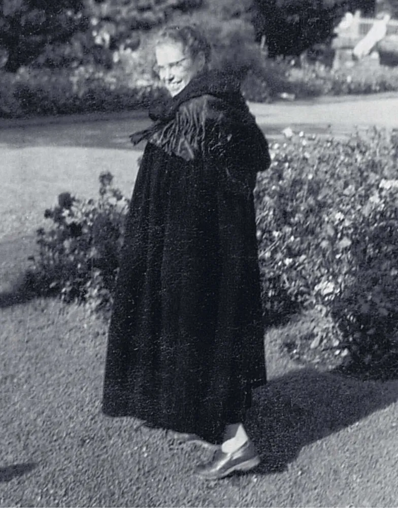 Schwarz-Weiß-Foto einer Frau in einem Garten. Sie trägt einen Mantel, eine Brille und lächselt.