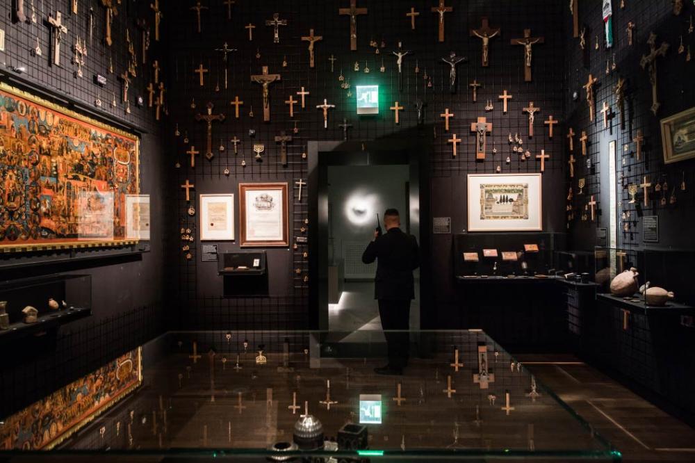 Interior view of an exhibition room full of crucifixes. A man with a walkie-talkie leaving the room is seen from behind. 