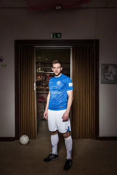 Ein junger Mann in Fu&szlig;ball-Outfit blickt in die Kamera. Links hinter ihm liegt ein Fu&szlig;ball. 