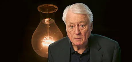 A white-haired man in the foreground, with a giant light bulb in the background.