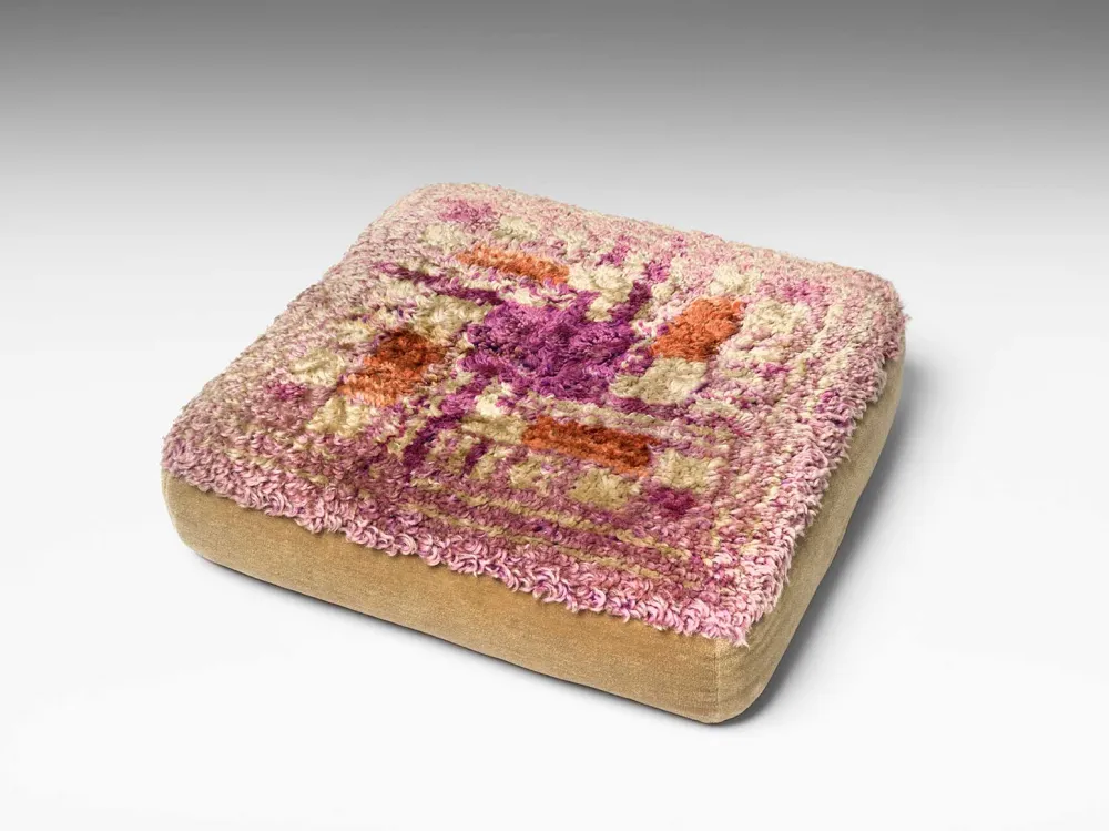 A beige-colored seat cushion that is knotted with pink, purple and white wool and features a graphic pattern.