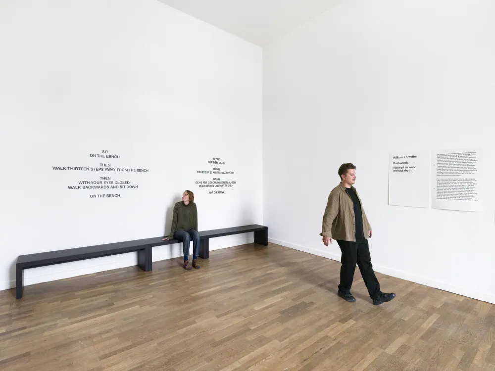 Eine Frau sitzt auf einer Bank. Ein Mann l&auml;uft r&uuml;ckwarts. Beide befinden sich in einem gro&szlig;en hellen Ausstellungsraum. 