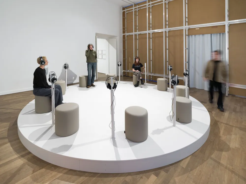 Mehrere Meschen befinden sich in einem gro&szlig;en Ausstellungsraum. Einige sitzen oder stehen auf einer Sitz-Insel im Raum und haben Kopfh&ouml;rer auf und h&ouml;ren Aufnahmen.