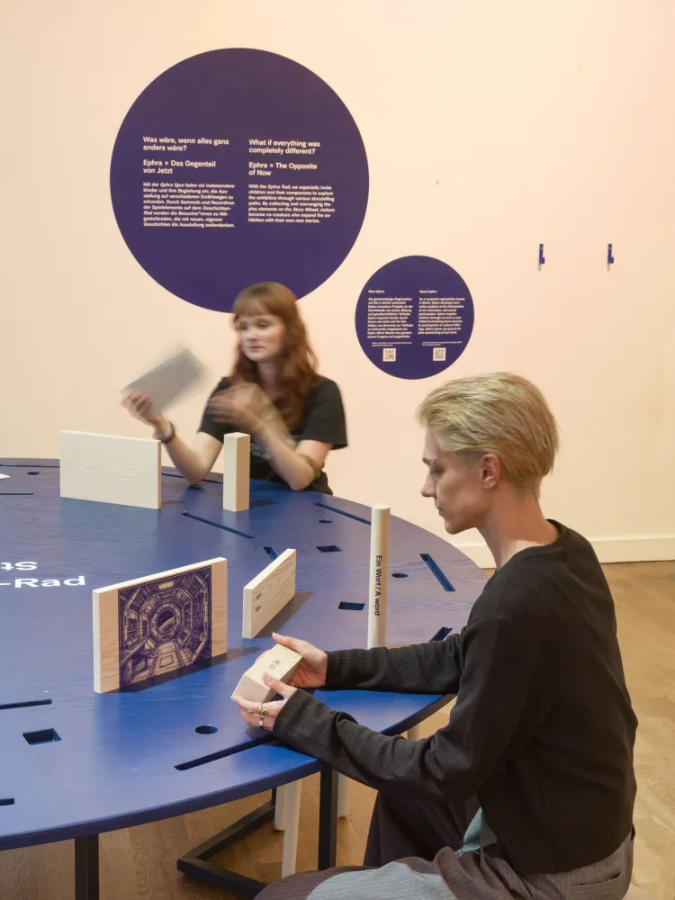 Zwei Menschen sitzen einem runden Tisch in einem Ausstellungsraum und schauen sich Objekte aus Holz an. 