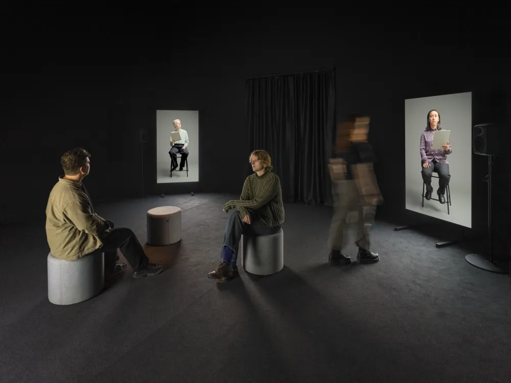 Zwei Menschen sitzen sich gegen&uuml;ber in einem dunklen Ausstellungsraum. Eine Person l&auml;uft durch den Raum. An den Wandseiten des Raumes sind zwei gro&szlig;e Monitore auf denen Menschen zu sehen sind, die auf St&uuml;hlen sitzen. 