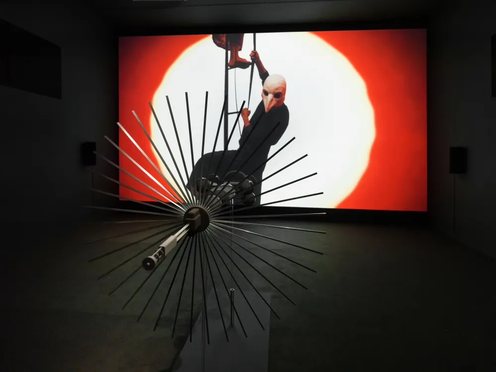 Futuristischer Metallgegenstand, der aussieht wie ein Ventilator, zeigt auf eine gro&szlig;e Filmleinwand. Auf der Leinwand steigen Personen eine Leiter empor. Eine Person hat eine Vogelkopfmaske auf. 