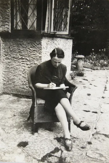  Black and white photograph of a woman sitting in a wicker chair on a terrace with a pen in her hand and a clipboard on her knees. She is looking at the board.