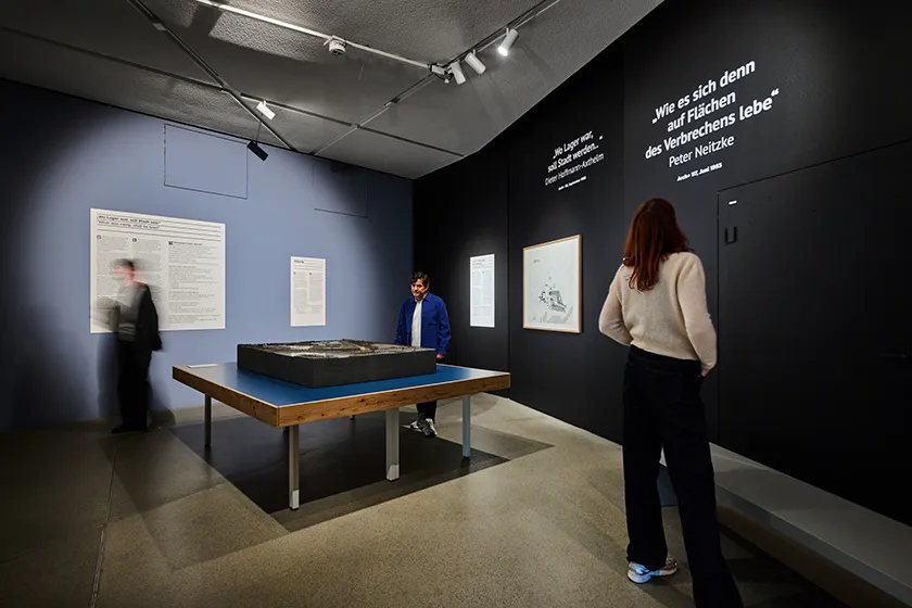Exhibition view with thee people either walking, observing objects or reading texts.
