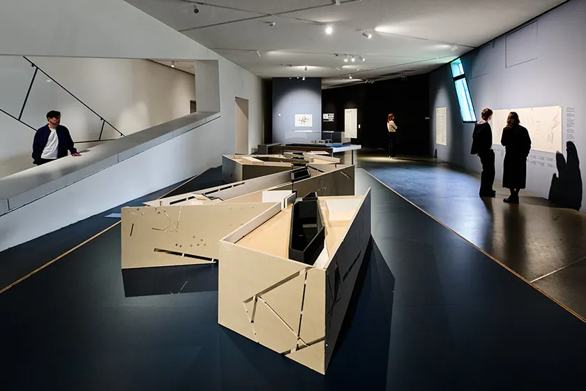 Exhibition view with three people: Two are standing in front of a wall reading a text, one is gazing into the space; the center part of the image and room itself is dominated by a zig-zag-shaped piece of furniture..