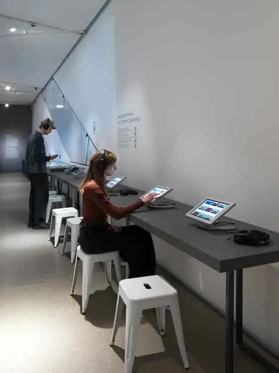 Das Bild zeigt einen Ausstellungsraum. Auf der rechten Seite an der Wand befinden sich schwarze Tische, auf denen Tablets angebracht sind. Vor den Tischen steht ein Mensch und auf einem der weißen Hocker sitzt eine Person.