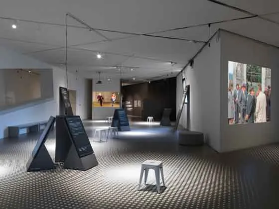 Das Bild zeigt einen Ausstellungsraum, in dem weiße Hocker und Aufsteller mit Text verteilt sind. Außerdem sieht man Projektionen von Videos auf Wänden.