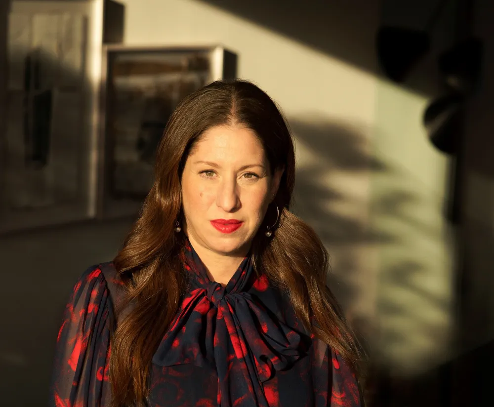 The picture shows a woman from the waist up. She is wearing a black and red dress and red lipstick and is looking straight at the camera.