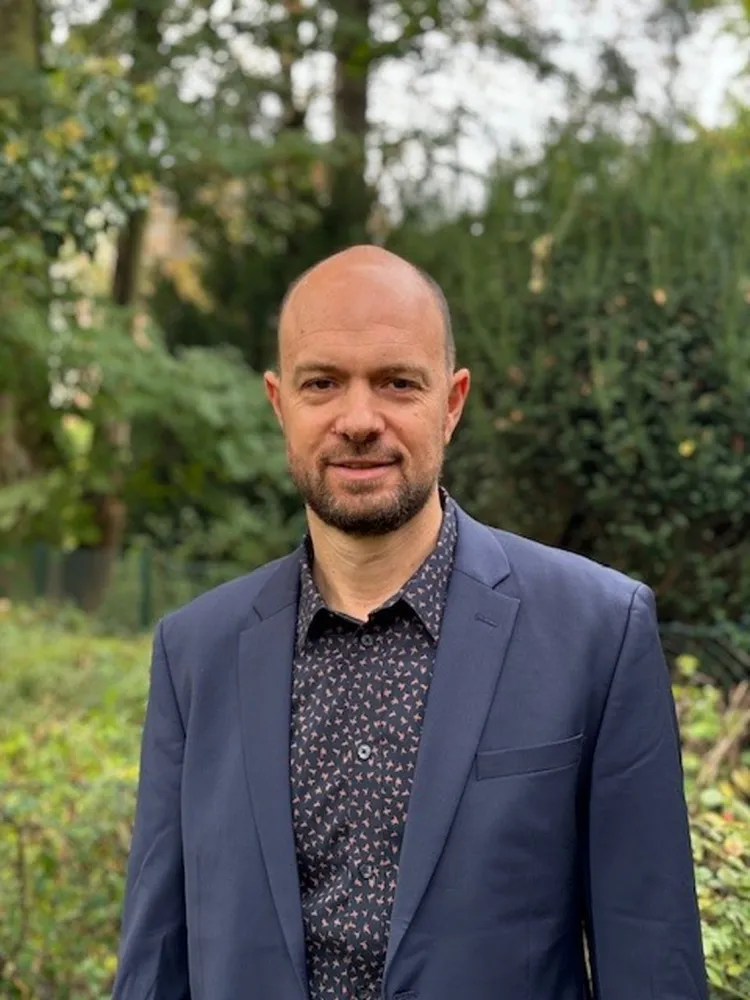 Mann schaut freundlich in die Kamera. Er tr&auml;gt ein blaues Jackett und ein gemustertes Hemd.