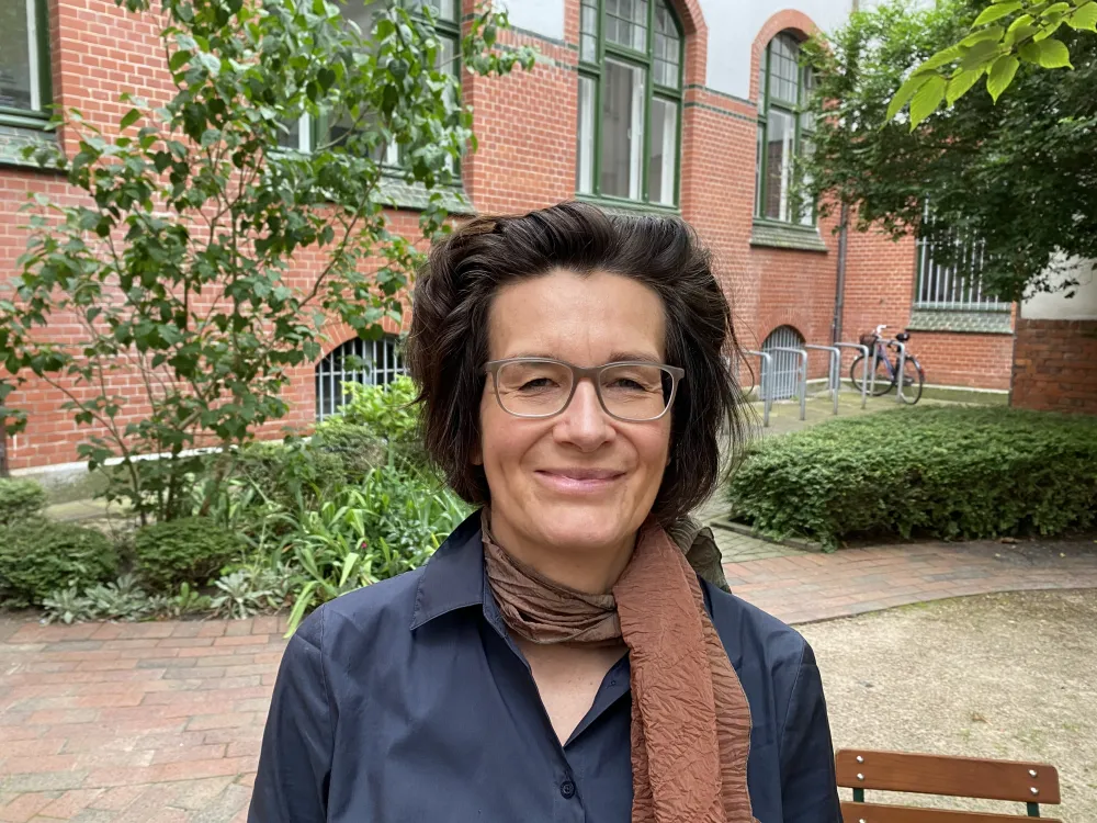 Portrait of a woman with dark hair and glasses. A building can be seen in the background. 