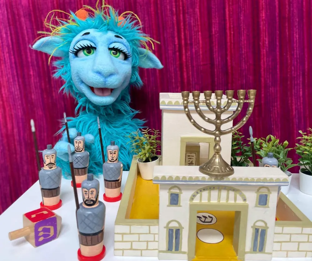 Eine blaue zottelige sprechende Puppe mit großen schönen Augen hinter einem Tisch mit Spielzeugfestung, Dreidel und Chanukka-Leuchter darauf.