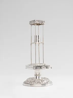 Silver ceremonial object: A small pedestal sits on a base, from which four struts rise upward, topped by a square piece that forms a roof-like structure. A candle holder is attached in the center.