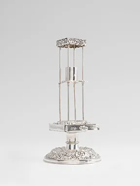 Silver ceremonial object: A small pedestal sits on a base, from which four struts rise upward, topped by a square piece that forms a roof-like structure. A candle holder is attached in the center.