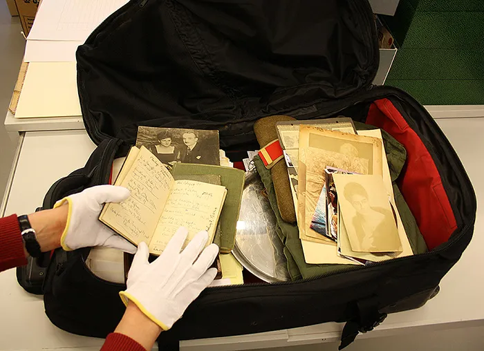 A briefcase containing various documents, photos, and other items. A person's hands are visible. They are wearing archival gloves and are taking out a small book.