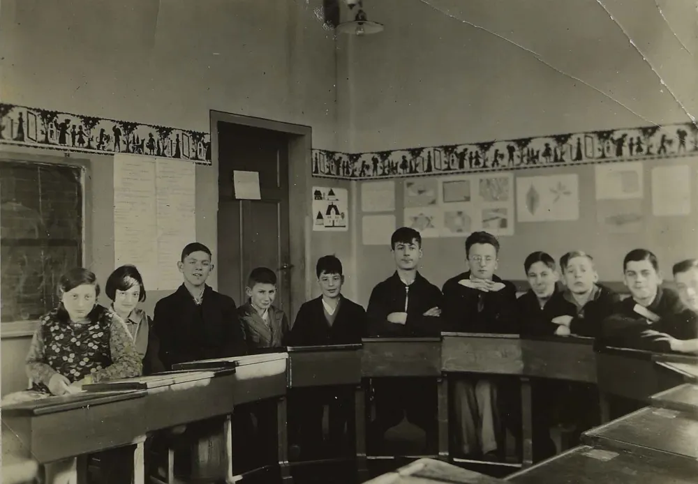 Das Bild zeigt einen Schulklassenraum mit elf Sch&uuml;lerinnen und Sch&uuml;lern, die in einem Halbkreis angeordnet an ihren Pulten sitzen.