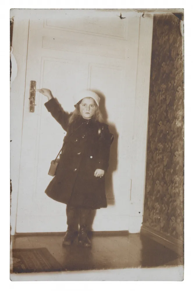 A little girl is standing in front of a door, holding the handle with one hand.