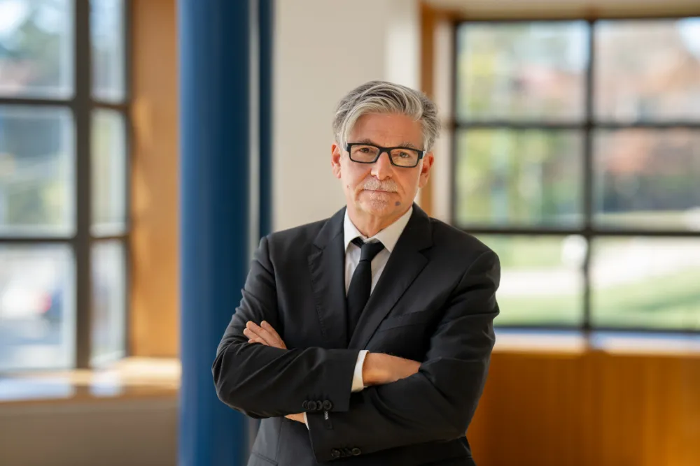 Mann mit Brille schaut freundlich in die Kamera. Er hat einen Anzug an und verschr&auml;nkt die Arme.