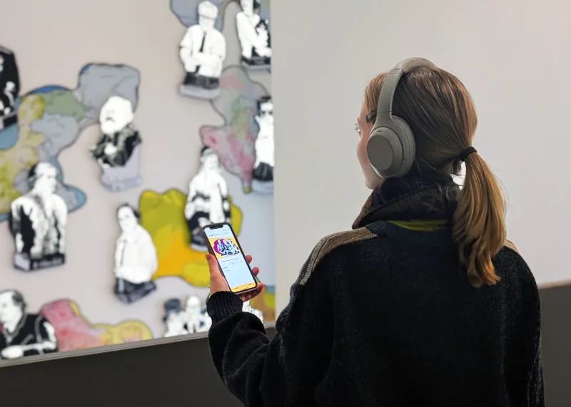 Portrait of a woman with long blonde hair and headphones in the Shabbat theme room. She is holding her mobile phone and following the JMB app Young Perspectives.