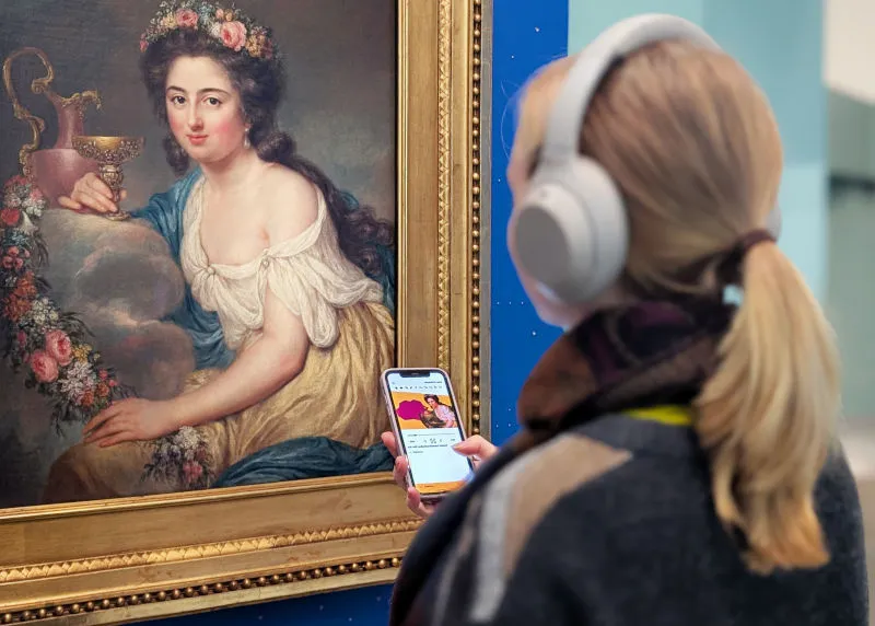 Portrait of a woman with long blonde hair and headphones in the Shabbat theme room. She is holding her mobile phone and following the JMB app Young Perspectives.