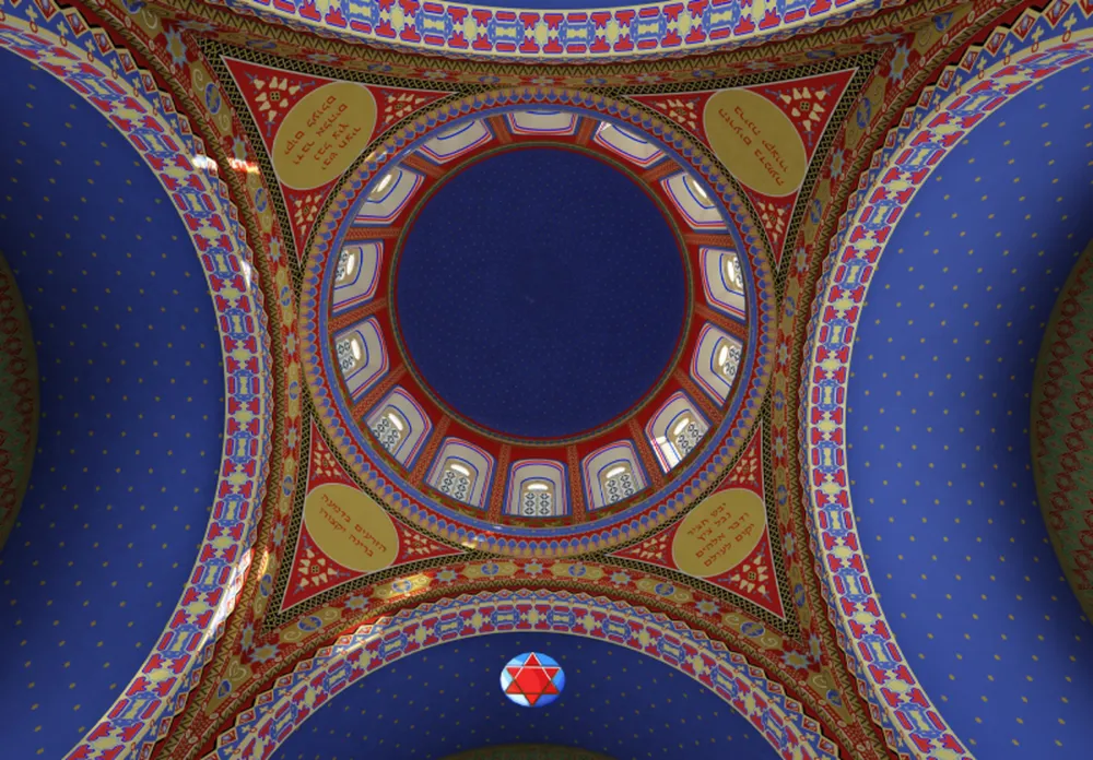 The picture shows the interior of a synagogue dome. Most of the ceiling is blue, and the various ornaments are yellow and red.