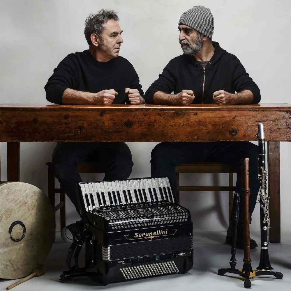 Two men are sitting next to each other at a wooden table, looking at each other, their four fists clenched on the tabletop. In front of the table are five instruments: a drum, an accordion, two flutes and a clarinet.