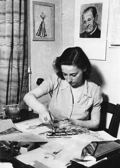  Black and white photo of a woman sitting at a table and dipping a brush in paint.