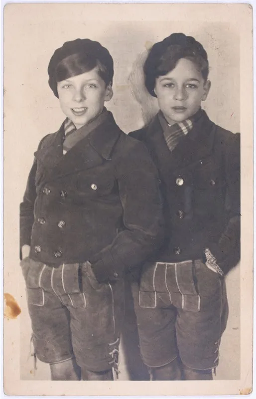 Black-and-white photo. Two boys wearing jackets and shorts stand next to each other and look at the camera.