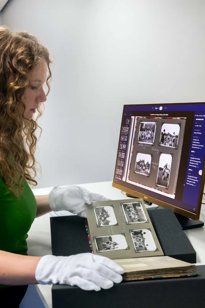 Eine Frau betrachtet ein historisches Fotoalbum und vergleicht es mit der Abbildung an einem Monitor. Sie trägt weiße Baumwollhandschuhe.