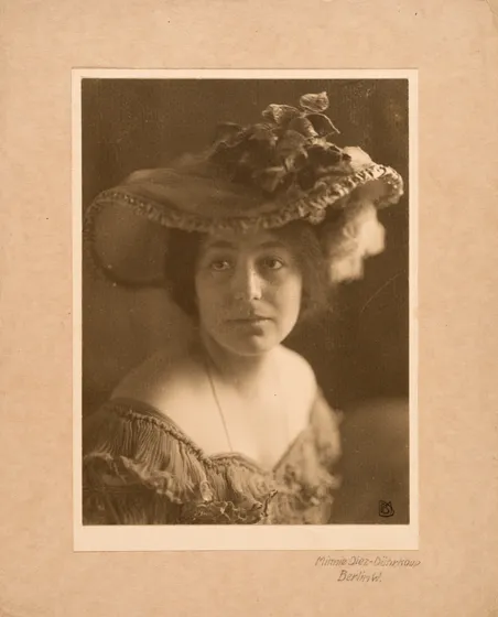  Sepia-colored portrait of a woman wearing a large, decorated hat and a dress with a boat neckline.