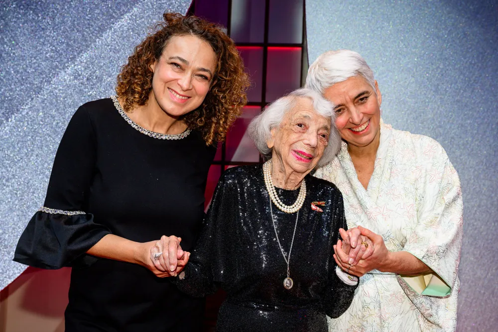 Three women are standing on a carpet, holding hands and smile.