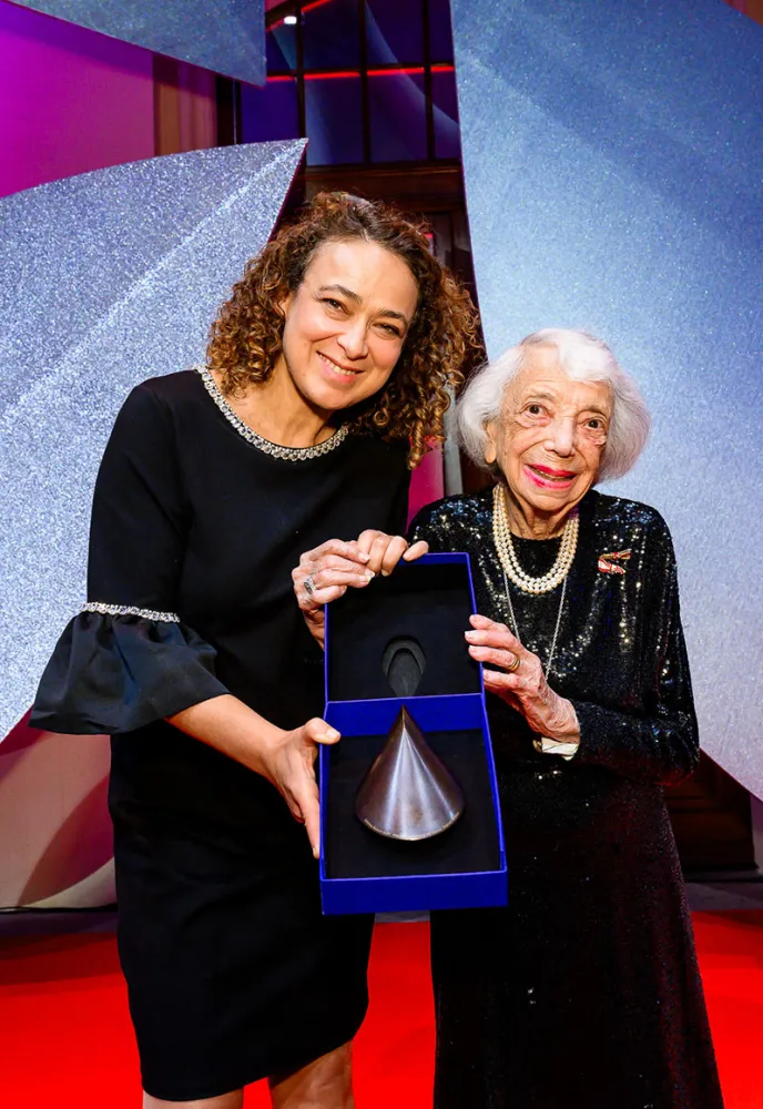 Two women stand on a red carpet holding their prize.