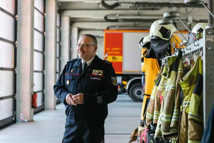 Ein Mann in Feuerwehruniform steht in einer Halle, rechts neben ihm an einer Garderobe h&auml;ngt Schutzkleidung.