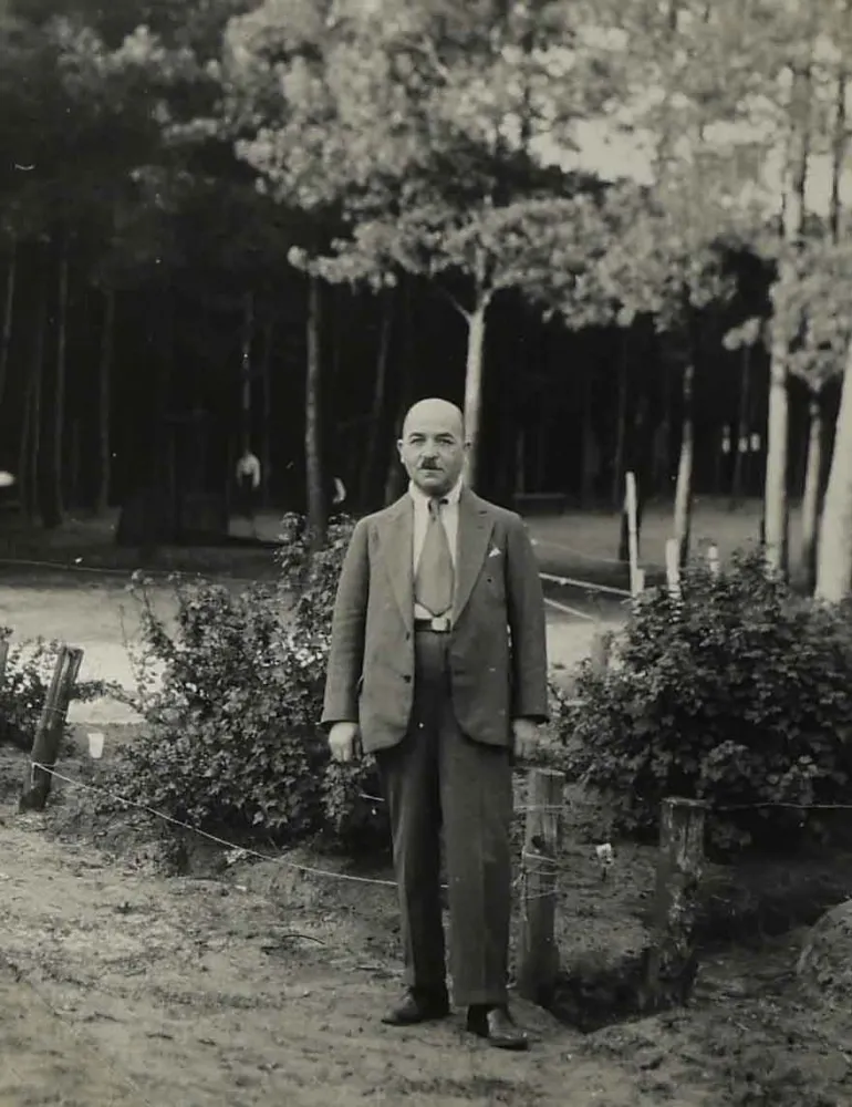 Das Bild zeigt einen Anzug tragenden Mann im Ganzk&ouml;rperportr&auml;t. Hinter ihm B&uuml;sche und B&auml;ume.