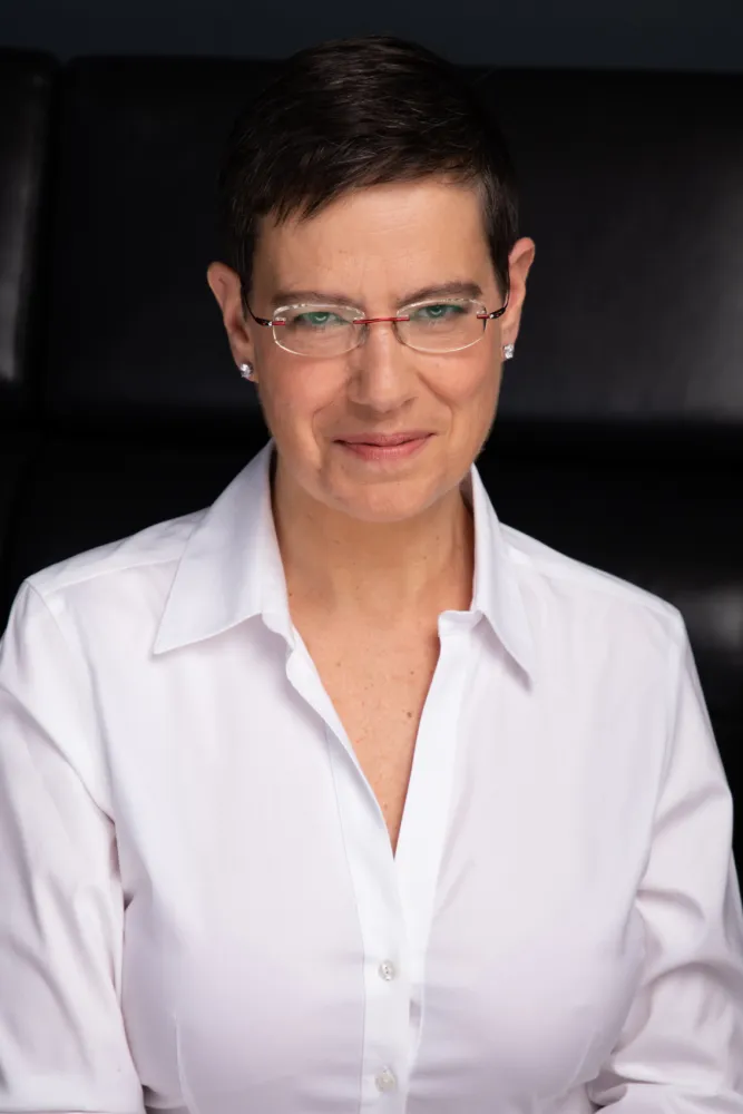 A woman with glasses and a white blouse looks kindly at the camera.