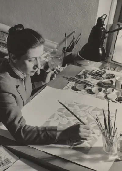 A woman is sitting at her desk covered with colours and brushes. She is painting.