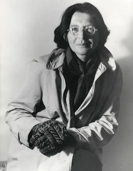  Black and white photograph of a woman wearing a light-colored coat and black gloves, resting on her raised knee.